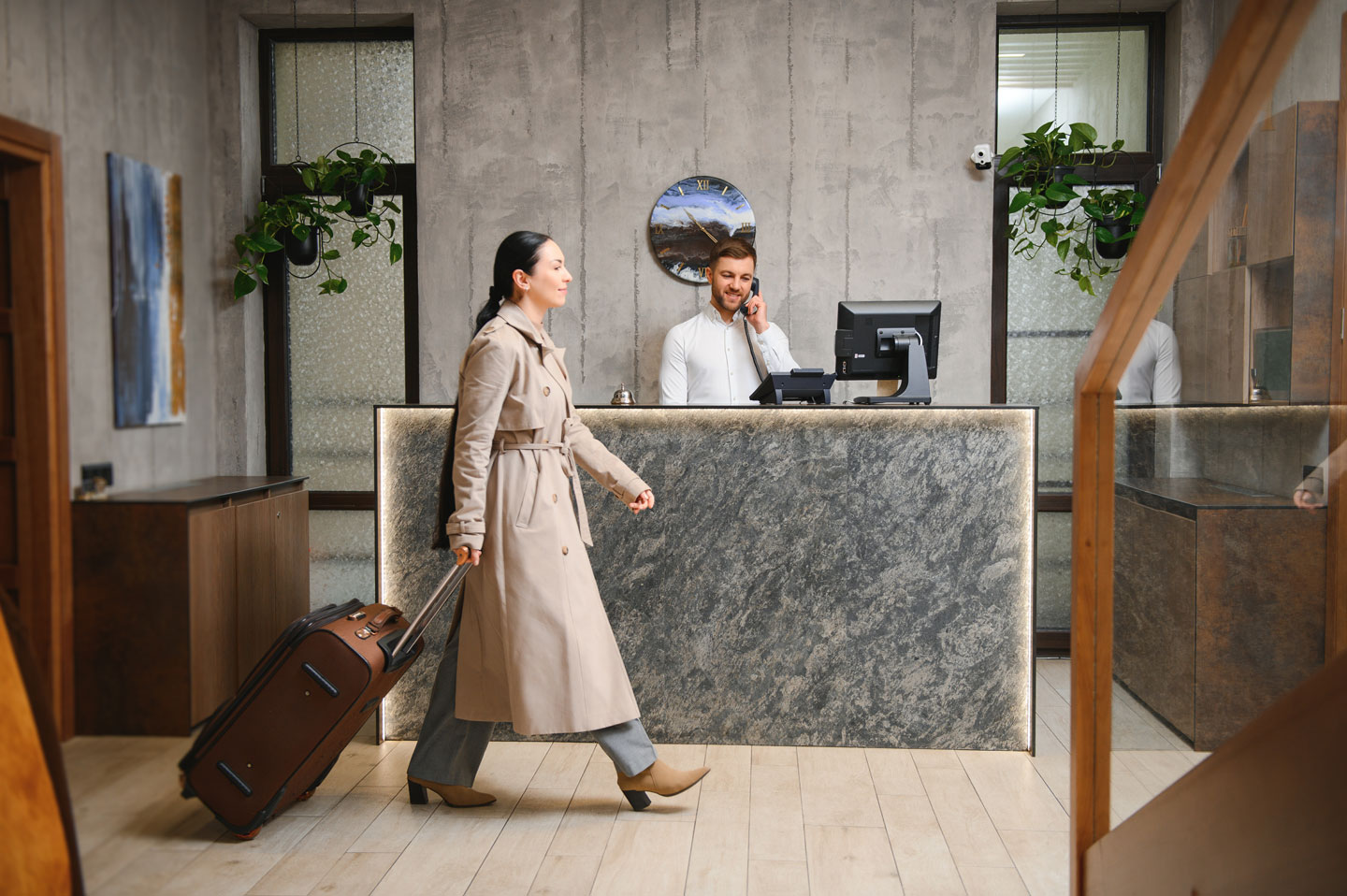 A hotel guest crosses the lobby with her travel case while a receptionist checks her in at the front desk.